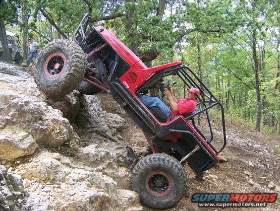 Red Jeep climbing a rocky hill with a person driving. Another off-road vehicle is visible in the background