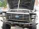 1986 Ford Bronco - Front view of a modified Ford Bronco, showing its grille and open hood. Another vehicle is partially visible to the left.