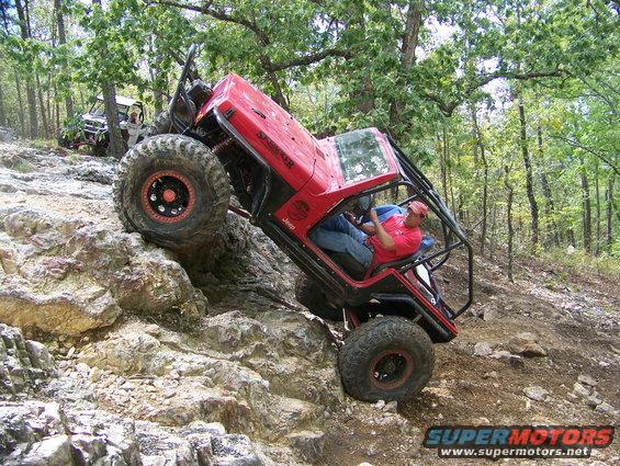 Red off-road Jeep tilted on rocks with a driver, another off-road vehicle behind
