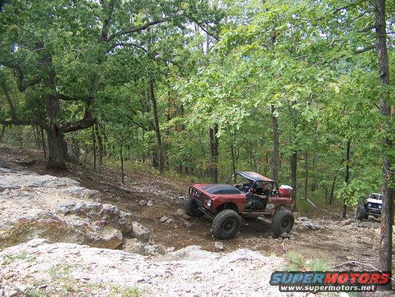 Red off-road vehicle with large tires on a rocky trail, another white vehicle behind it