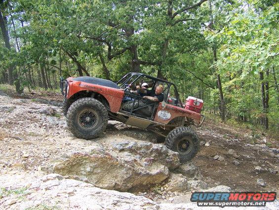 Custom off-road buggy with roll cage climbing over rocks in a wooded area