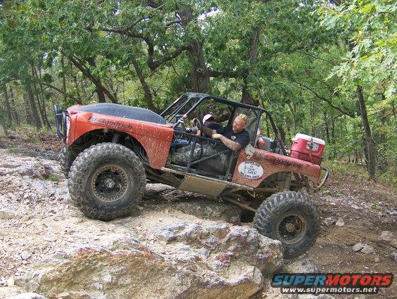 Orange off-road buggy with a roll cage driving over rocks in a wooded area. Driver has a beard and is wearing a black shirt