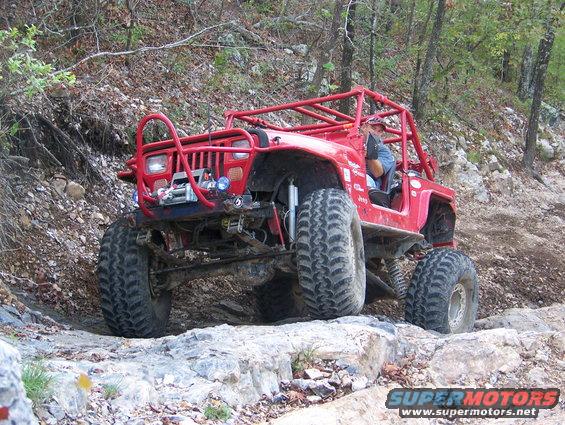 Red Jeep with roll cage and large tires driving on a rocky trail