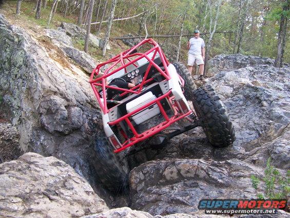 Off-road buggy with a red roll cage going over rocks, a man stands nearby