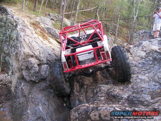 Red roll cage buggy with large tires climbing rocks, rear view
