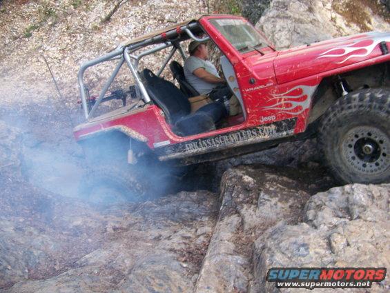 Red Jeep Wrangler with flame decals and a roll cage going over rocks, kicking up smoke