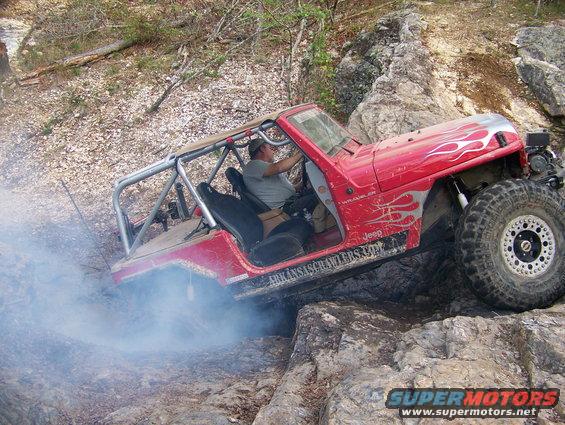 Red Jeep Wrangler with flame decals and roll cage navigating rocky terrain, emitting smoke from the rear tire