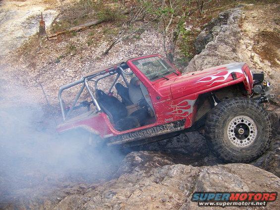 Red Jeep Wrangler with flame decals navigating rocky terrain, emitting smoke