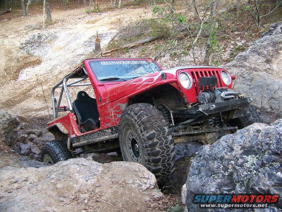 Red Jeep Wrangler with custom roll cage and winch navigating a rocky trail. "ARKANSASCLAWLERS.COM" visible on windshield