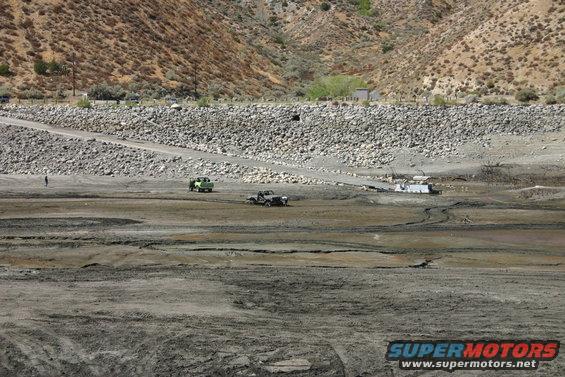 Two off-road vehicles traverse a dry, muddy terrain with a rocky hillside in the background