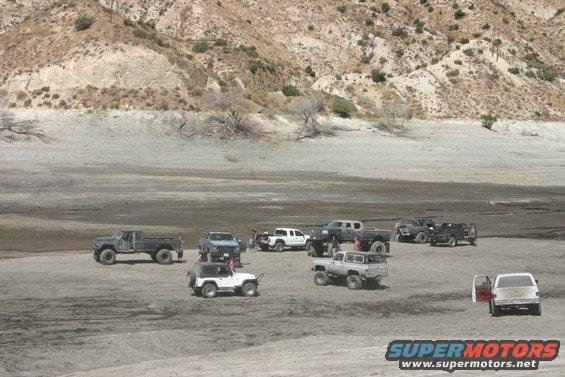 Several off-road trucks and SUVs parked in a dry, dusty landscape with hills in the background. One white SUV has its doors open