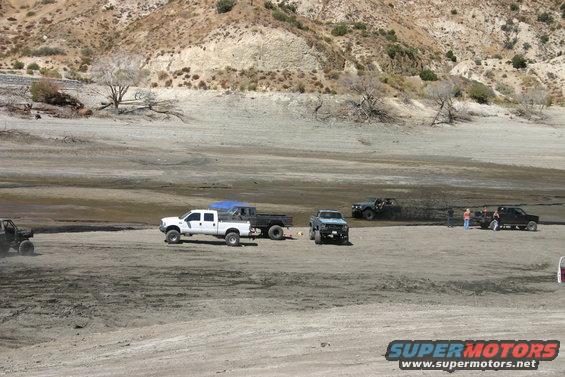 Several trucks and off-road vehicles gathered in a dry, dusty area with hills in the background