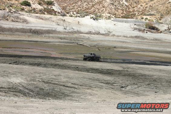 Dark-colored 1980s Ford Bronco driving on a dry, dusty plain with sparse vegetation and hills in the background