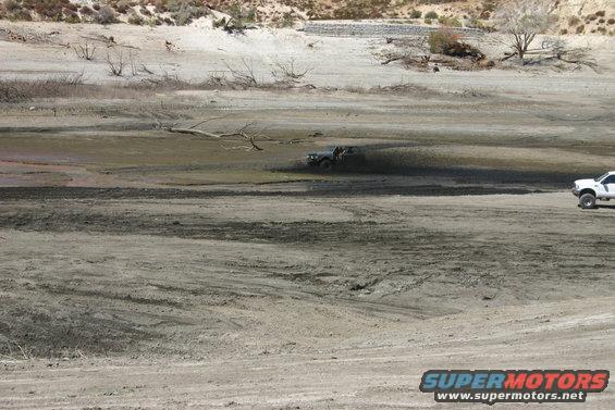Dark off-road vehicle kicking up mud in dry lake bed; white pickup truck parked to the right