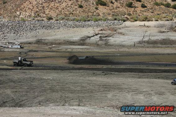Two trucks driving through mud, one kicking up a large spray