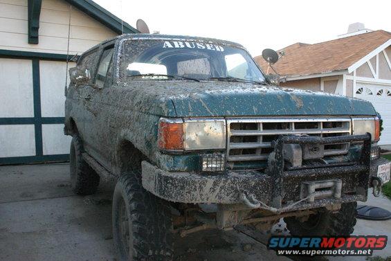 Mud-covered 1991 Ford Bronco parked in front of a garage, with "ABUSED" sticker on windshield