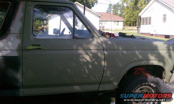 Olive green Ford Bronco side view, showing door, window, and tire. Some parts are disassembled