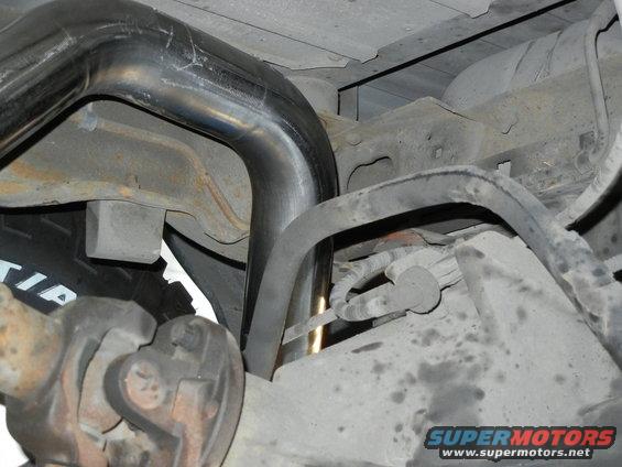 Underside of a vehicle showing a large exhaust pipe curving over the rear axle and suspension components. Tire sidewall is partially visible