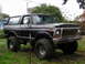 1979 Ford Bronco - This black Ford Bronco is parked on grassy terrain with trees and structures in the background.