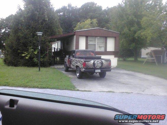 Pickup truck with camouflage and winged graphic on tailgate, parked in driveway of mobile home
