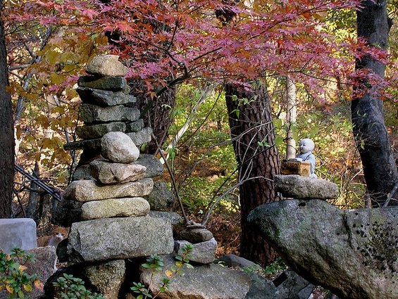 Stone cairns and a small statue sit among trees with autumn leaves