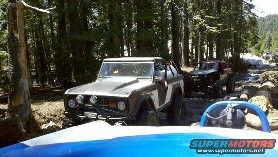 Brown and white Ford Bronco off-roading with a red truck behind it