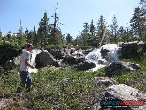 Man walking near a waterfall in a forest with large rocks and green bushes