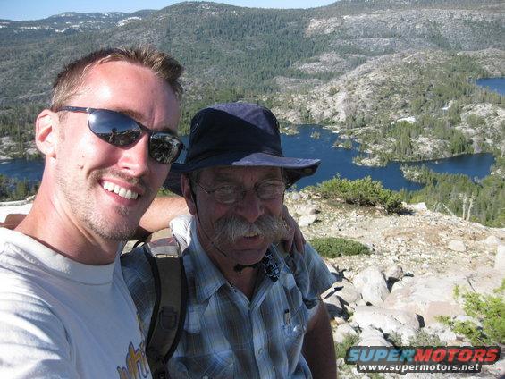 Two men smile for a selfie with a scenic mountain lake landscape behind them