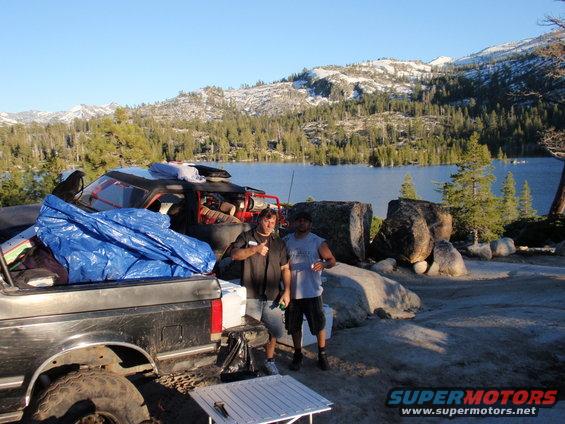 Two men stand by a pickup truck loaded with camping gear next to a lake with snow-capped mountains in the background