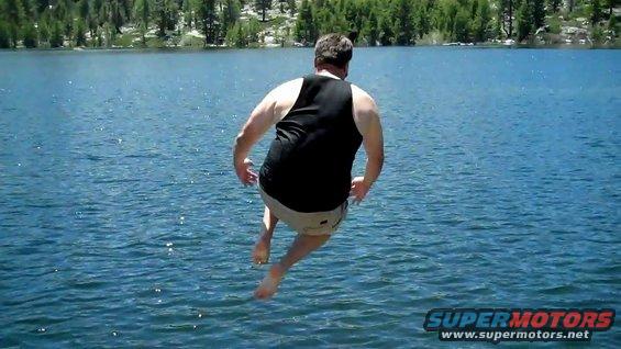 Man in black tank top and shorts jumps into a blue lake with trees and mountains in background. Supermotors.net logo in lower right