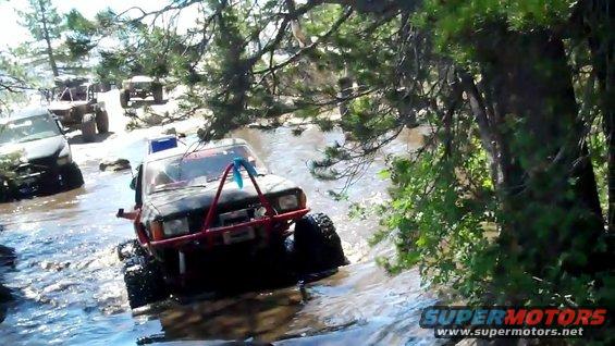 Several off-road vehicles driving through a shallow river with trees on the right
