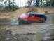 1979 Ford Bronco - A red Jeep Wrangler Rubicon navigates a muddy off-road trail, kicking up water and mud.