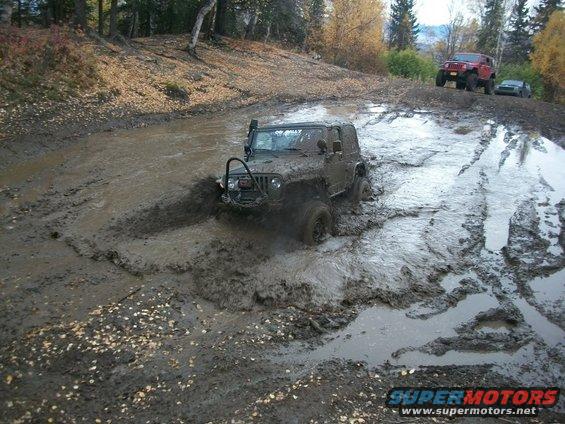 Mud-covered Jeep Wrangler splashing through a muddy track, with a red Jeep and a dark car in the background