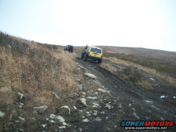 Two SUVs driving off-road on a muddy, rocky hill under a cloudy sky