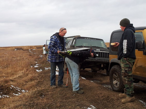 Two men are working on a black Jeep Cherokee, another yellow Jeep is parked nearby. A third person stands in the background