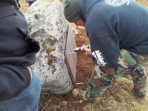 Two people are drilling into a large rock next to a wooden panel. One person wears a dark hoodie and camouflage pants