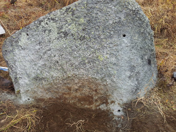 Big rock with several holes drilled into it, sitting in dry grass and dirt