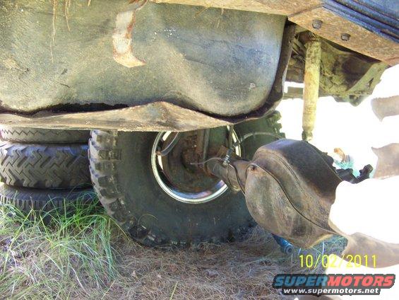 Underside of a vehicle showing the fuel tank, rear axle, and a shock absorber. Tires are stacked nearby