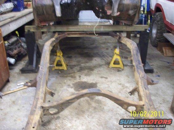 Vehicle frame supported by jack stands in a garage. A partial vehicle body is visible above the frame
