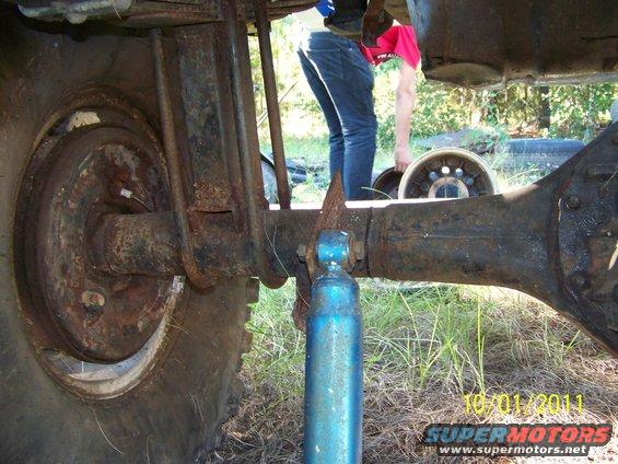 close-up of rusty rear axle, leaf springs, and blue shock absorber on a vehicle; person in background holding a wheel rim