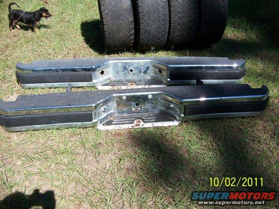 Two chrome and black truck bumpers lying on grass, with tires and a dog in the background