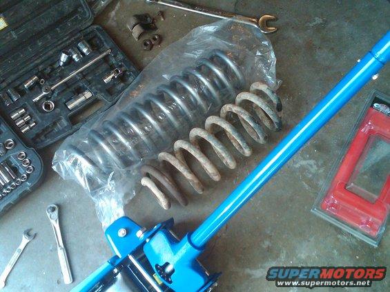 Two suspension springs, one new and one rusty, next to a blue jack and a toolbox