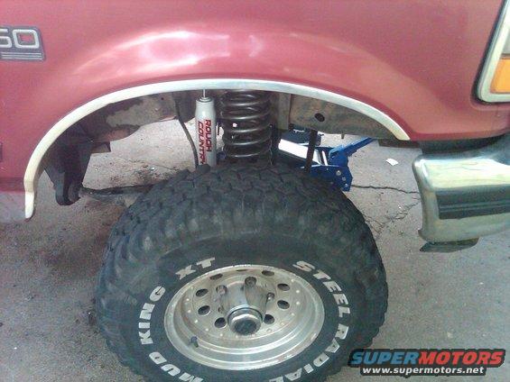 Close-up of a red Ford F-Series truck's front wheel well, showing a large mud tire, a Rough Country shock, and a blue jack under the frame