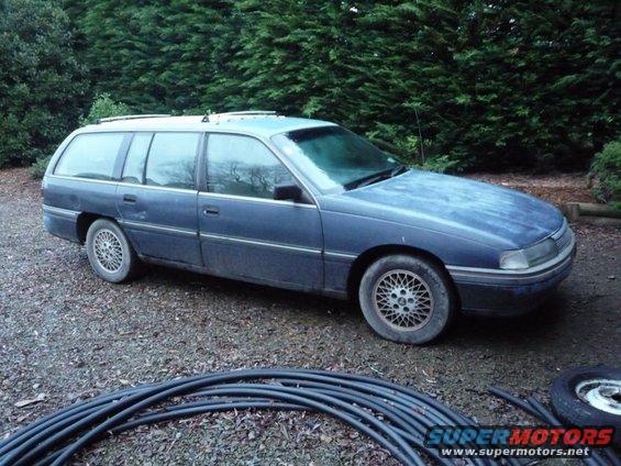 Blue station wagon with roof racks parked on gravel, trees in background