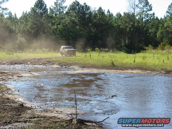 1994 Ford Bronco kicking up dust driving through a muddy field near trees