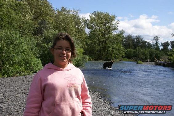 Girl in pink hoodie stands on rocky shore with bear wading in river behind her