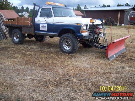 1973 Ford F-250 flatbed truck with a red plow attachment, white and blue cab, yellow light bar
