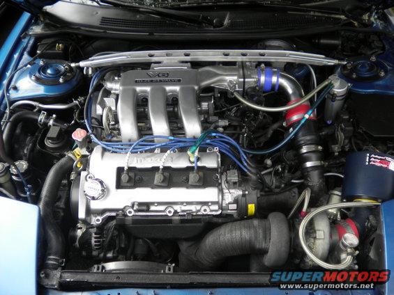 Engine bay of a blue 1995 Ford Probe, showing a V6 engine with aftermarket turbocharger and intake. Visible "Mazda" and "V6" badges