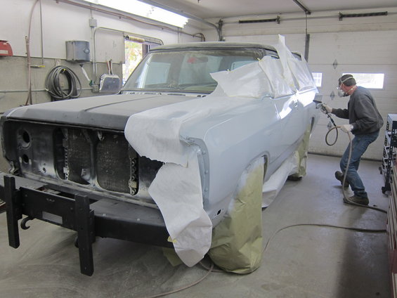 1987 Dodge 1500 pickup truck in a body shop, being prepped for paint. Hood is black, body is primer gray. Man sprays paint on fender
