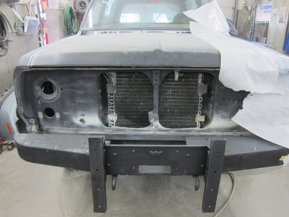 Truck front end, primer gray, missing headlights and grille, with a black aftermarket bumper
